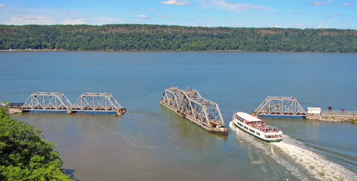 A Circle Line sightseeing boat passing the open Spuyten Duyvil swing bridge from the Harlem River side into the Hudson, with the Palisades behind, seen on a New York architecture boat tour